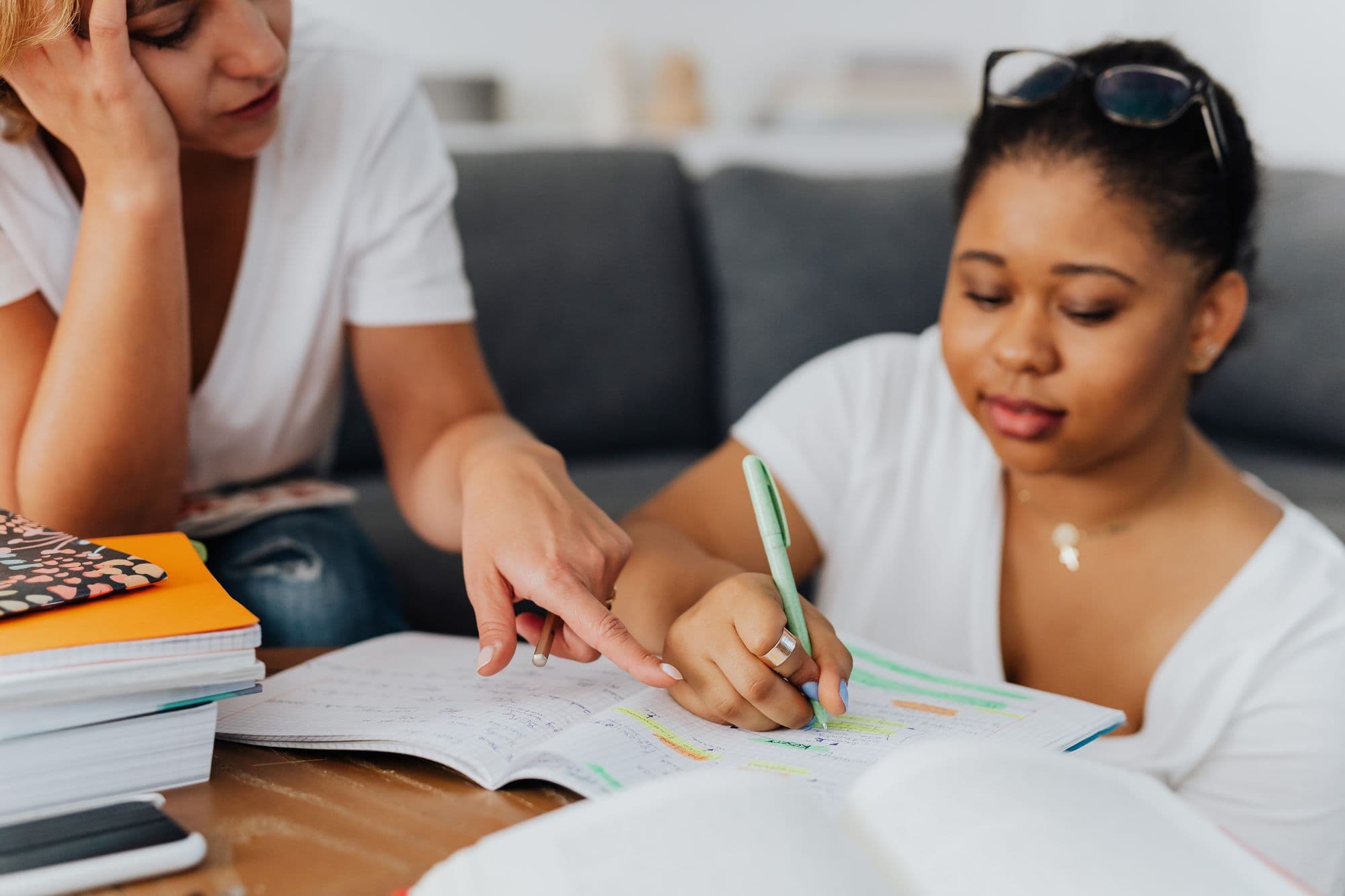 Uma professora aponta uma anotação no caderno enquanto a aluna escreve, na sala de casa.